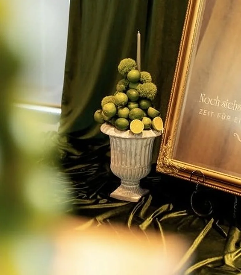 Weiße Urne mit Limetten vor goldgerahmter Willkommenstafel